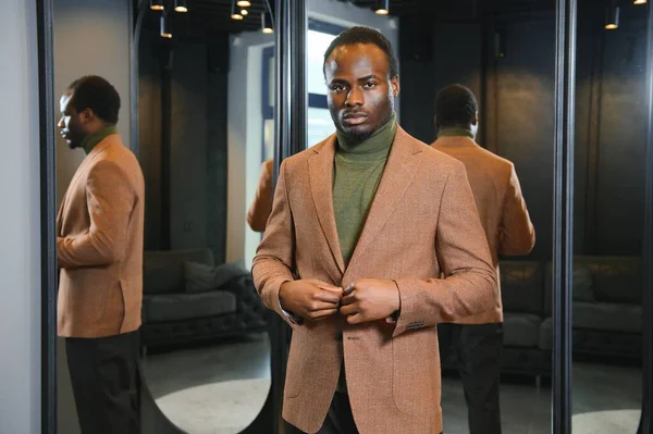 İndirim, alışveriş, moda, tarz ve insan konsepti - Afrikalı bir adam ceketini ya da takım elbisesini seçiyor ve alışveriş merkezinde ya da giyim mağazasında aynaya bakıyor.