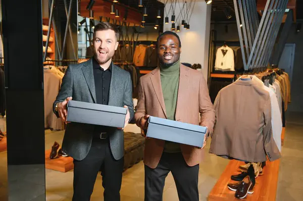 Giysi kutuları taşıyan iki mutlu çok ırklı adam bir giyim mağazasında duruyor..