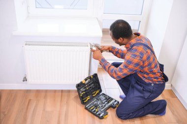 Çalışma kıyafetli bir adam ısıtma kaloriferini yerleştirirken veya tamir ederken araçlar kullanıyor.