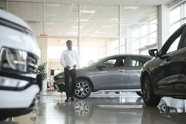 Cars Dealership Concept. Auto Seller Afro Man Standing In Automobile Center.