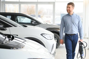 Yakışıklı bir iş adamı elektrikli araba için kablolu yayın tutuyor. Beyaz erkek galeride elektrikli otomobilin yanında duruyor..