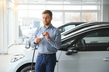 Yakışıklı bir iş adamı elektrikli araba için kablolu yayın tutuyor. Beyaz erkek galeride elektrikli otomobilin yanında duruyor..
