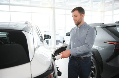 Yakışıklı bir iş adamı elektrikli araba için kablolu yayın tutuyor. Beyaz erkek galeride elektrikli otomobilin yanında duruyor..