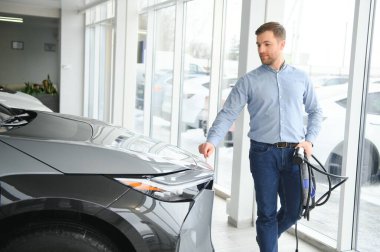 Yakışıklı bir iş adamı elektrikli araba için kablolu yayın tutuyor. Beyaz erkek galeride elektrikli otomobilin yanında duruyor..