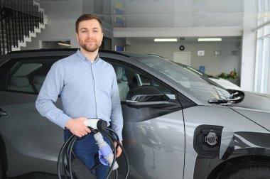 Yakışıklı bir iş adamı elektrikli araba için kablolu yayın tutuyor. Beyaz erkek galeride elektrikli otomobilin yanında duruyor..