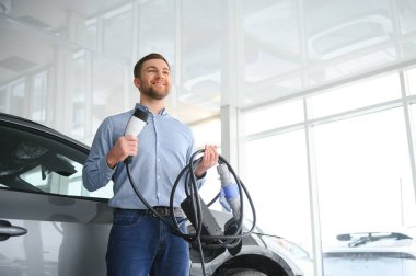 Yakışıklı bir iş adamı elektrikli araba için kablolu yayın tutuyor. Beyaz erkek galeride elektrikli otomobilin yanında duruyor..