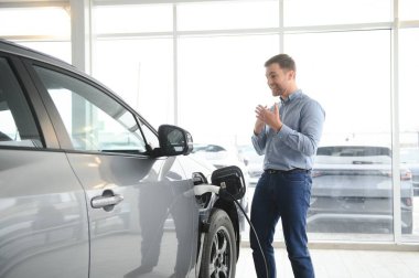 Yakışıklı bir iş adamı elektrikli araba için kablolu yayın tutuyor. Beyaz erkek galeride elektrikli otomobilin yanında duruyor..