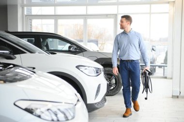 Yakışıklı bir iş adamı elektrikli araba için kablolu yayın tutuyor. Beyaz erkek galeride elektrikli otomobilin yanında duruyor..