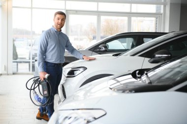 Yakışıklı bir iş adamı elektrikli araba için kablolu yayın tutuyor. Beyaz erkek galeride elektrikli otomobilin yanında duruyor..