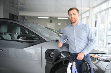 Yakışıklı bir iş adamı elektrikli araba için kablolu yayın tutuyor. Beyaz erkek galeride elektrikli otomobilin yanında duruyor..