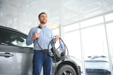 Elektrikli araba alma kavramı. Yakışıklı iş adamı galeride elektrikli arabanın yanında duruyor..