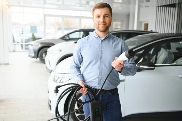 Elektrikli araba alma kavramı. Yakışıklı iş adamı galeride elektrikli arabanın yanında duruyor..