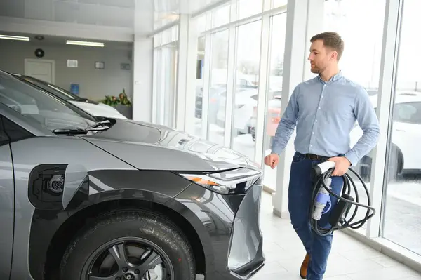 Elektrikli araba alma kavramı. Yakışıklı iş adamı galeride elektrikli arabanın yanında duruyor..