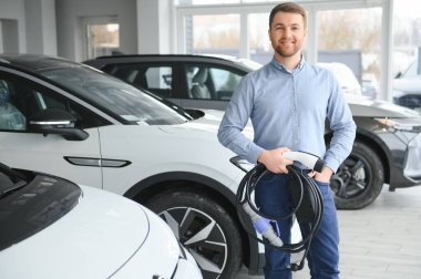 Yakışıklı bir iş adamı elektrikli araba için kablolu yayın tutuyor. Beyaz erkek galeride elektrikli otomobilin yanında duruyor..