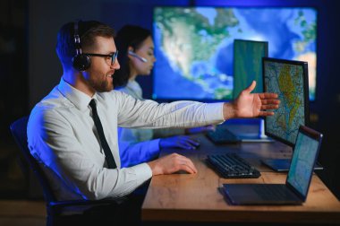 Oda izleme içinde teknik destek uzmanı kulaklık konuşuyor. Onun meslektaşları arka planda çalışan.