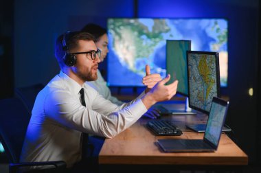 Oda izleme içinde teknik destek uzmanı kulaklık konuşuyor. Onun meslektaşları arka planda çalışan.