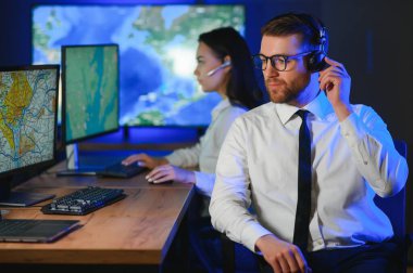 Oda izleme içinde teknik destek uzmanı kulaklık konuşuyor. Onun meslektaşları arka planda çalışan.