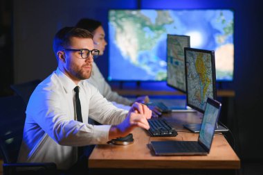 Oda izleme içinde teknik destek uzmanı kulaklık konuşuyor. Onun meslektaşları arka planda çalışan.