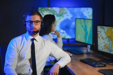 Oda izleme içinde teknik destek uzmanı kulaklık konuşuyor. Onun meslektaşları arka planda çalışan.