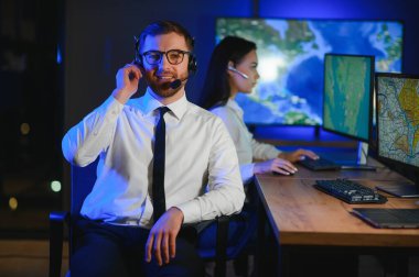 Oda izleme içinde teknik destek uzmanı kulaklık konuşuyor. Onun meslektaşları arka planda çalışan.