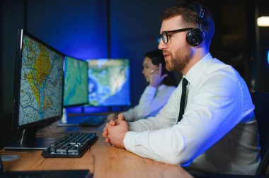 Oda izleme içinde teknik destek uzmanı kulaklık konuşuyor. Onun meslektaşları arka planda çalışan.