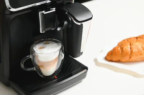 Modern coffee machine with glass cup of latte on white marble countertop in kitchen.