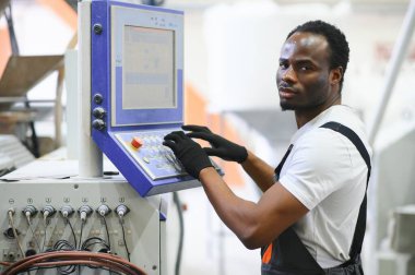 Afrikalı erkek mühendis, şirketin ürün işiyle ilgili plastik ve çelik endüstrisinde çalışarak, malzeme üretim makinesi sistemini kontrol etmek için makine dairesinde duruyor..