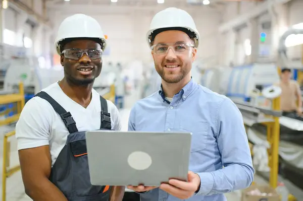 Two Diverse Professional Heavy Industry Engineers Wearing Safety Uniform and Hard Hats Working on Laptop.