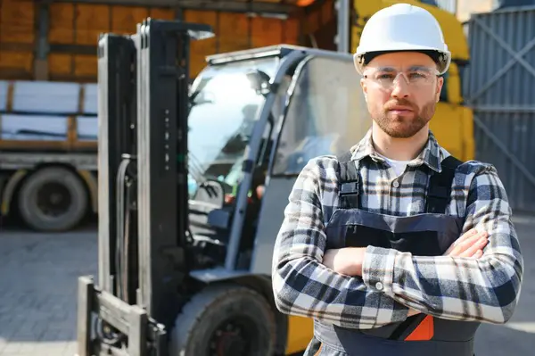 forklift sürücüsü koruyucu yelek ve forklift yük nakliye şirketi deposunda ayakta, gülümseyerek.
