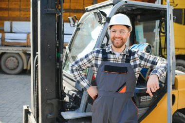 forklift sürücüsü koruyucu yelek ve forklift yük nakliye şirketi deposunda ayakta, gülümseyerek.