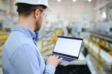 Endüstriyel fabrikada duran kulaklık ve dizüstü bilgisayarlı teknisyen veya mühendisin yan görüntüsü.