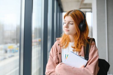 Kızıl saçlı öğrenci kız elinde kitaplarla pencerenin önünde duruyor..