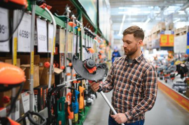 Bahçe malzemeleri dükkanındaki genç bir adam çim biçmek için elektrikli budama makinesi seçiyor..
