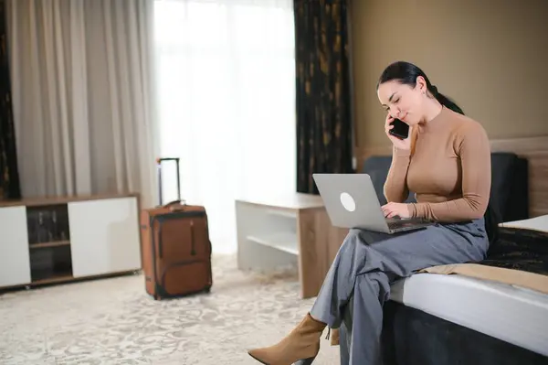 brunette white businesswoman sitting on bed in a hotel room talking on mobile phone using laptop computer.