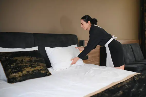 Young pretty housekeeper in uniform changing bedclothes in hotel room while standing by double bed and putting clean cover