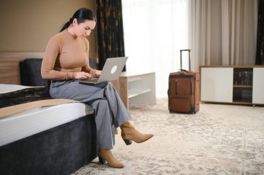 İş gezisinde otel odasından çalışan genç bir iş kadını, yatakta oturan ve dizüstü bilgisayar kullanan bir kadın..