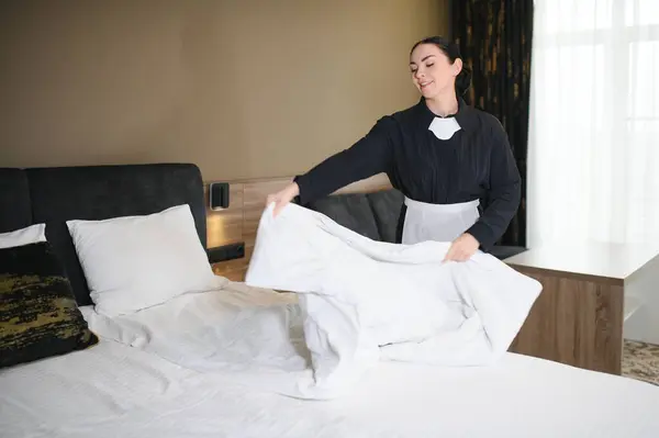 Young pretty housekeeper in uniform changing bedclothes in hotel room while standing by double bed and putting clean cover