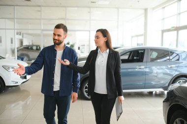 Gülümseyen araba satıcısı müşteriyle birlikte araba galerisinde duruyor ve satılık arabaları gezdiriyor..