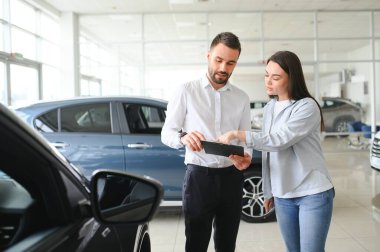 Gülümseyen araba satıcısı müşteriyle birlikte araba galerisinde duruyor ve satılık arabaları gezdiriyor..