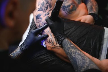 Dövme tabancası tutan erkek dövme sanatçısı, dövmeli bir erkek modelin koluna dövme yapma sürecini gösteriyor..