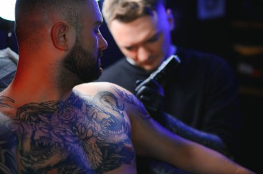 Dövme tabancası tutan erkek dövme sanatçısı, dövmeli bir erkek modelin koluna dövme yapma sürecini gösteriyor..