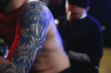 Dövme tabancası tutan erkek dövme sanatçısı, dövmeli bir erkek modelin koluna dövme yapma sürecini gösteriyor..