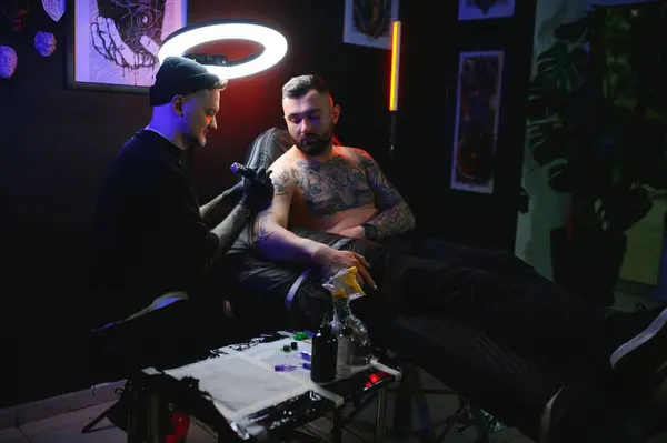bearded tattoo artist working at his studio tattooing sleeve on the arm of his male client. Man getting tattooed by professional tattooist.