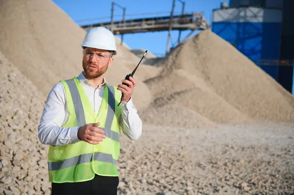 A worker in a hard hat stands with a walkie-talkie at a stone crushing plant