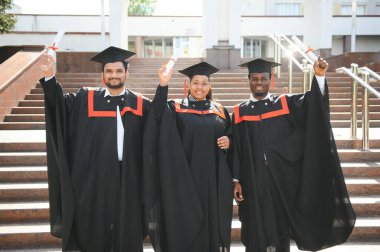 Mezuniyet elbiseleri ve şapkaları içinde, elinde diplomalarıyla mutlu, çok kültürlü bir grup insan kampüste kameraya bakarak üniversiteden mezun oluyor. Üniversite mezunu, üniversite konsepti.