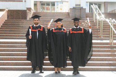 Mezuniyet törenine katılan çeşitli uluslararası öğrenciler mezuniyet şapkası ve cüppeli mutlu çok ırklı öğrenciler üniversite kampüsünde sıraya dizilmiş.