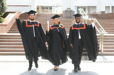 Mezuniyet elbiseleri ve şapkaları içinde, elinde diplomalarıyla mutlu, çok kültürlü bir grup insan kampüste kameraya bakarak üniversiteden mezun oluyor. Üniversite mezunu, üniversite konsepti.