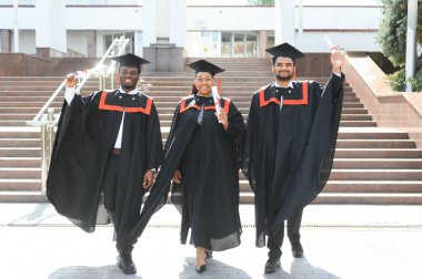 Mezuniyet törenine katılan çeşitli uluslararası öğrenciler mezuniyet şapkası ve cüppeli mutlu çok ırklı öğrenciler üniversite kampüsünde sıraya dizilmiş.