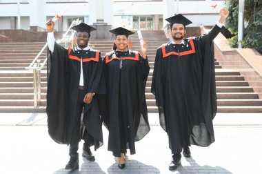 Mezuniyet elbiseleri ve şapkaları içinde, elinde diplomalarıyla mutlu, çok kültürlü bir grup insan kampüste kameraya bakarak üniversiteden mezun oluyor. Üniversite mezunu, üniversite konsepti.