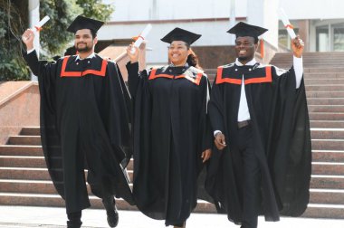 Mezuniyet elbiseleri ve şapkaları içinde, elinde diplomalarıyla mutlu, çok kültürlü bir grup insan kampüste kameraya bakarak üniversiteden mezun oluyor. Üniversite mezunu, üniversite konsepti.